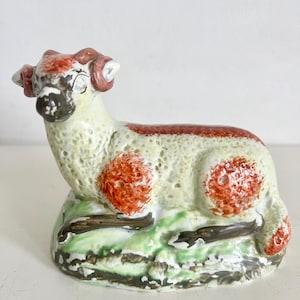 May include: A ceramic sheep figurine with a cream-colored body, red accents, and a green base. The sheep has red horns and spots, and is resting on a base with green grass and brown accents. The figurine is a decorative item.