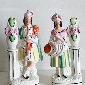 Puede incluir: Un par de figuras de porcelana antiguas. Las figuras visten trajes de época, una con un abanico y la otra con un haz. Cada figura se alza sobre un pedestal blanco con un elemento decorativo. La paleta de colores incluye blanco, amarillo, verde, rosa y naranja.