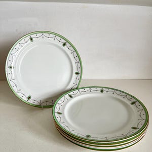 May include: White ceramic plates with a green and brown decorative border. The plates have a simple design with a green rim and a pattern of small dots and leaf-like shapes. A stack of plates is visible.