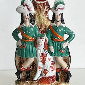 Könnte beinhalten: Vintage Keramikvase mit zwei Figuren in grünen Mänteln und Hüten mit orangefarbenen Schärpen, die einen weiß-rot gefleckten Hund flankieren. Die Figuren stehen zu beiden Seiten eines Baumstamms mit einer roten Öffnung.