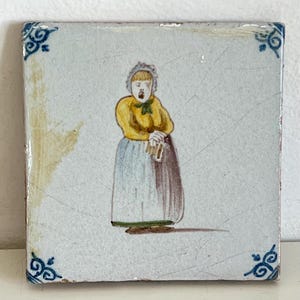 May include: Square ceramic tile featuring a hand-painted illustration of a person in a yellow top and blue skirt. The tile has blue corner designs and a distressed appearance, suggesting age.