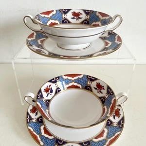 May include: Two white porcelain soup bowls with matching saucers. The bowls have two handles and are decorated with a blue, red, and gold floral pattern. The saucers have a matching design. The set is displayed on a clear surface.