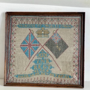 May include: A framed textile artwork with two crossed flags, a crown, and text. The flags are diamond-shaped, one with a Union Jack design and the other with a green and black pattern. The artwork is in a brown wooden frame.