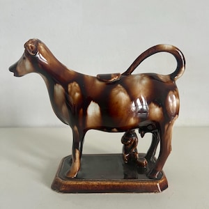 Pode incluir: Um cremeira de vaca de cerâmica marrom com uma alça curva e uma pequena figura na base. A vaca está em uma base retangular, e a cobertura tem um efeito de gotejamento.