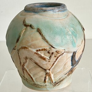 Può includere: Un vaso in ceramica fatto a mano con un corpo arrotondato e un bordo leggermente svasato. Il vaso presenta una combinazione di colori azzurro e beige con disegni lineari marroni e strutturati. Il bordo è blu più scuro.