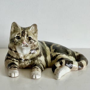 Może przedstawiać: Ceramiczna figurka kota w pozycji leżącej, o błyszczącym wykończeniu z brązowymi i czarnymi pręgami na kremowym tle. Kot ma bursztynowe oczy, różowy nos i białe łapy, a jego ogon jest owinięty wokół boku ciała.