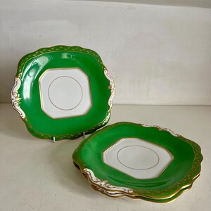 Op de afbeelding: Twee groene en witte porseleinen borden met gouden rand. De borden hebben een geschulpte rand en een vierkant midden met een gouden ring. Eén bord staat rechtop en de andere is gestapeld met nog twee borden.