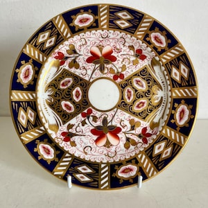 May include: A decorative plate with a white centre and a detailed border. The border features a navy blue and gold geometric design with red floral accents. The plate is supported by two white stands.