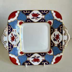 May include: Square serving plate with a white center and a decorative border. The border features a blue, red, and white floral pattern with gold trim. The plate has handles on each side.