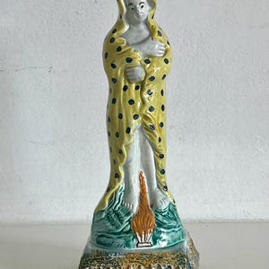 Könnte beinhalten: Keramikfigur einer stehenden Figur, die in einen gelben Umhang mit schwarzen Punkten gehüllt ist. Die Figur hat einen hellen Teint und steht auf einem grün-braunen Sockel mit einer Flamme.