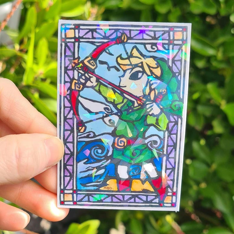 May include: A stained glass style sticker featuring a cartoon character holding a bow and arrow. The character is wearing a green tunic and a green hat. The sticker has a rainbow iridescent finish.