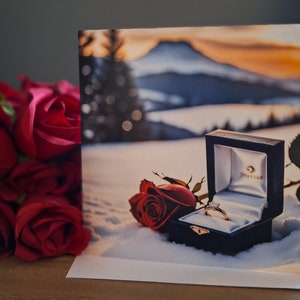 May include: A white card with a photograph of a ring in a black box with a red rose on top. The background is a snowy mountain scene with a sunset.