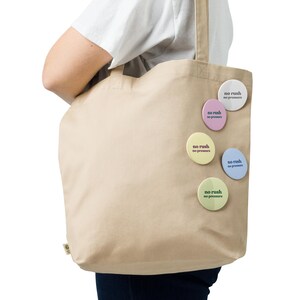 May include: A beige canvas tote bag with several round, pastel-coloured buttons attached. Each button has the text "no rush no pressure" in a simple font. The bag is held by a person.