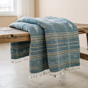 May include: A blue woven throw blanket featuring a grid pattern of white and olive green horizontal and vertical stripes. The fabric is adorned with small, embroidered diamond-shaped motifs and finished with short, knotted fringe along the edges.