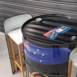 Puede incluir: Una mesa de barril de metal negro con la bandera australiana, un logotipo de Penrite y el texto Celebrating Years Australian Made & Owned Since 1926. El barril tiene un acabado brillante con dos tapones de metal y una pegatina rectangular.