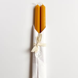 May include: Two beeswax candles wrapped in white tissue paper with a brown ribbon and a white label with the text "SWOYE" and a small tree design.