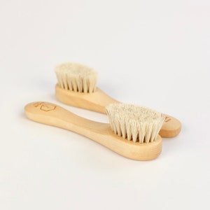 Può includere: Due spazzole per il viso in legno con setole di colore chiaro sono esposte su uno sfondo bianco. Le spazzole hanno un manico curvo e una testina rotonda. Il legno è di colore chiaro naturale. Le setole sono fitte e sembrano morbide.