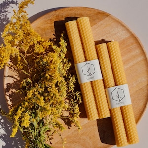May include: Three natural beeswax candles on a wooden plate with a sprig of yellow flowers. The candles are labeled "Swoye" and have a honeycomb pattern.