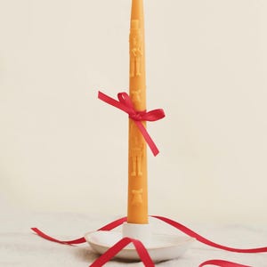 Pode incluir: Uma vela laranja acesa com figuras em relevo, amarrada com uma fita vermelha, está em um castiçal de cerâmica branca. A fita vermelha contorna a base. Uma decoração festiva.