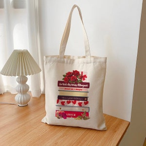 Könnte beinhalten: Eine weiße Canvas-Tragetasche mit einem Design aus einem Stapel Bücher mit roten Rosen und dem Text "La Nuit des temps, R. Barjavel" und "Avant toi, J. Moyes". Das unterste Buch hat das Wort "AMOUR" darauf gedruckt.