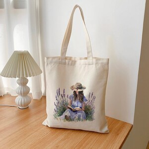 Peut inclure: Sac fourre-tout crème avec une illustration à l'aquarelle d'une personne lisant dans un champ de lavande. La personne porte une robe bleue et un chapeau de paille. Le sac a de longues anses.