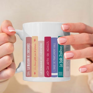 Könnte beinhalten: Eine weiße Keramiktasse mit einem Design aus gestapelten Büchern. Die Buchtitel sind in Französisch und beinhalten "La Maison de l'Orchidée", "La Jeune fille sur la falaise", "Le Domaine de l'Héritière", "La Rose de minuit", "L'Ange de Marchmont Hall", "La Belle Italienne" und "Le Secret de l'Enfer".