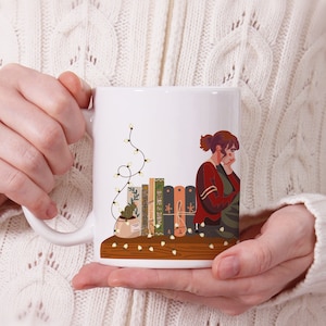 Könnte beinhalten: Weiße Keramiktasse mit einer bunten Illustration einer Person, die ein Buch liest, mit einer Lichterkette darüber. Die Bücher haben Titel wie "Geographie" und "Wein & Geruch".