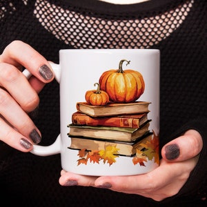Könnte beinhalten: Eine weiße Keramiktasse mit einem Herbstmotiv. Die Tasse zeigt einen Stapel Bücher mit zwei Kürbissen darauf. Die Kürbisse sind orange und braun mit grünen Stielen. Die Bücher sind braun und haben goldene Akzente. Um die Basis der Bücher herum befinden sich Herbstblätter.
