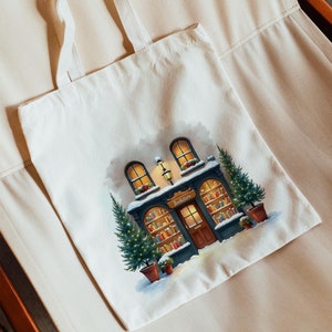 Könnte beinhalten: Eine weiße Canvas-Tragetasche mit einer winterlichen Szene eines Buchladens mit einem Schild, auf dem "Buchhandlung" steht. Der Buchladen hat eine Tür und Fenster mit eingeschalteten Lichtern. Vor dem Buchladen stehen zwei Weihnachtsbäume mit eingeschalteten Lichtern. Die Szene spielt vor einem verschneiten Hintergrund.