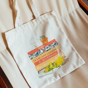 Könnte beinhalten: Cremefarbene Tragetasche mit literarischem Thema. Die Tasche zeigt einen Stapel Bücher mit Titeln wie "POESIE" und "LE LAC, A.DE LAMARTINE." Florale und botanische Illustrationen und Text sind auf die Tasche gedruckt.
