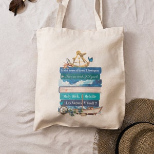 Könnte beinhalten: Eine weiße Canvas-Tragetasche mit einem Design aus einem Stapel Bücher mit nautischen Motiven. Die Bücher haben Titel in Französisch und Englisch, darunter "Le vieil homme et la mer", "Face au vent", "Moby Dick" und "Les Vagues".