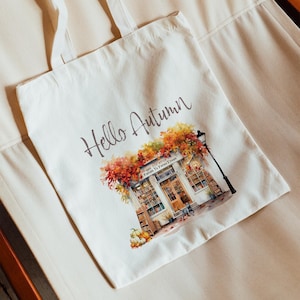 Könnte beinhalten: Eine weiße Stofftasche mit dem Aufdruck "Hello Autumn". Die Tasche zeigt eine Aquarell-Illustration eines Buchladens mit Herbstlaub und einer schwarzen Katze.