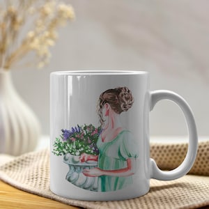 Könnte beinhalten: Weiße Keramiktasse mit einer Aquarell-Illustration einer Frau in einem grünen Kleid, die einen Blumenstrauß hält. Die Frau schaut nach links von der Tasse.
