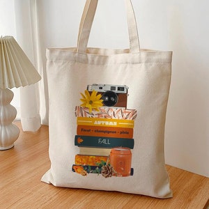 Könnte beinhalten: Eine weiße Canvas-Tragetasche mit einem Design aus einem Stapel Bücher, einer Kamera, einer Sonnenblume und einer Kerze. Die Bücher tragen die Worte "AUTUMN" und "FALL". Der Text auf den Büchern lautet "Foret - champignon - pluie".
