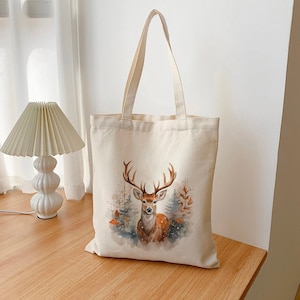 Peut inclure: Un sac fourre-tout crème avec une illustration à l'aquarelle d'un cerf avec de grands bois. Le cerf est placé sur un fond d'arbres et de feuilles d'automne. Le sac a deux longues poignées.