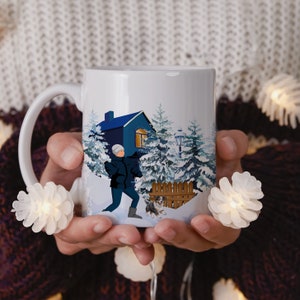 Könnte beinhalten: Weiße Keramiktasse mit einer Winterszene, die eine Person zeigt, die einen Hund vor einem blauen Haus mit schneebedeckten Bäumen spazieren führt.
