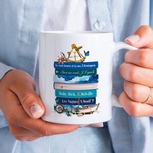 Könnte beinhalten: Weiße Keramiktasse mit einem blau-weißen nautischen Design. Die Tasse zeigt einen Kompass, ein Segelboot, Schmetterlinge und einen Stapel Bücher mit Titeln in Französisch und Englisch. Die Titel beinhalten "Le vieil homme et la mer", "Face au vent", "Moby Dick" und "Les Vagues".