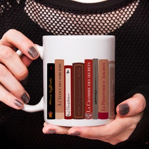 Könnte beinhalten: Weiße Keramiktasse mit einem Design aus einem Stapel Bücher mit Titeln in Französisch. Die Titel beinhalten "Maison Gryffender", "À L'ÉCOLE DES SORCIERS", "bus Dumbledore", "LA CHAMBRE DES SECRETS", "LE PRISONNIER D'AZKABAN" und "J. K. Rowling".