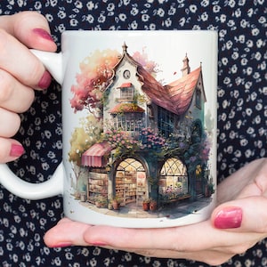 Könnte beinhalten: Weißer Keramikbecher mit einer Aquarellillustration eines malerischen Buchladens. Das Gebäude hat eine rote Markise, gewölbte Fenster und ein rotes Ziegeldach, umgeben von Blumen und Bäumen. Der Becher wird von einer Person gehalten.