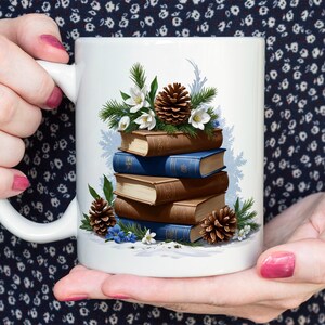 Peut inclure: Mug en céramique blanche orné d'une pile de livres aux couvertures marron et bleues, agrémentée de pommes de pin, de fleurs blanches et de verdure. Le design évoque un thème hivernal et chaleureux.