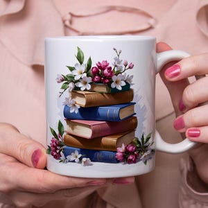 Peut inclure: Une tasse en céramique blanche avec une illustration colorée d'une pile de livres avec des fleurs roses et blanches. Les livres sont bleus, bruns, roses et blancs.