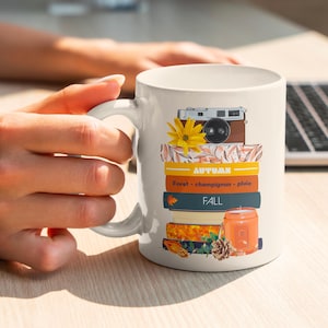 Könnte beinhalten: Weiße Keramiktasse mit einem Herbst-Motiv. Das Design zeigt einen Stapel Bücher mit einer Kamera, einer Sonnenblume und einer Kerze obenauf. Die Bücher sind mit den Worten "AUTUMN" und "FALL" beschriftet.