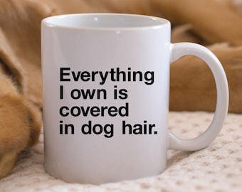 May include: White ceramic mug with the text "Everything I own is covered in dog hair" in black. The mug has a curved handle and is set against a blurred background.