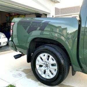 May include: A green pickup truck with a black "FRONTIER" decal on the side. The truck has a black tire and a silver rim. The truck is parked in a driveway.