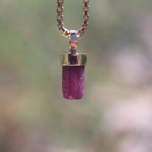 May include: A gold-toned necklace with a rectangular chain and a pendant. The pendant features a cylindrical, deep pink gemstone encased in a gold setting. The background is a soft, blurred green and brown.
