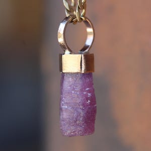 May include: A gold chain necklace with a pendant featuring a raw, purple gemstone. The pendant is attached to a gold ring and has a gold cap at the top.