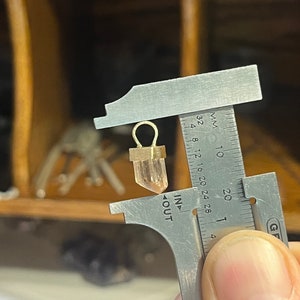 May include: A close-up of a clear quartz crystal pendant with a gold-toned setting. The pendant is being measured with a caliper, showing a measurement of approximately 1.5 centimeters.
