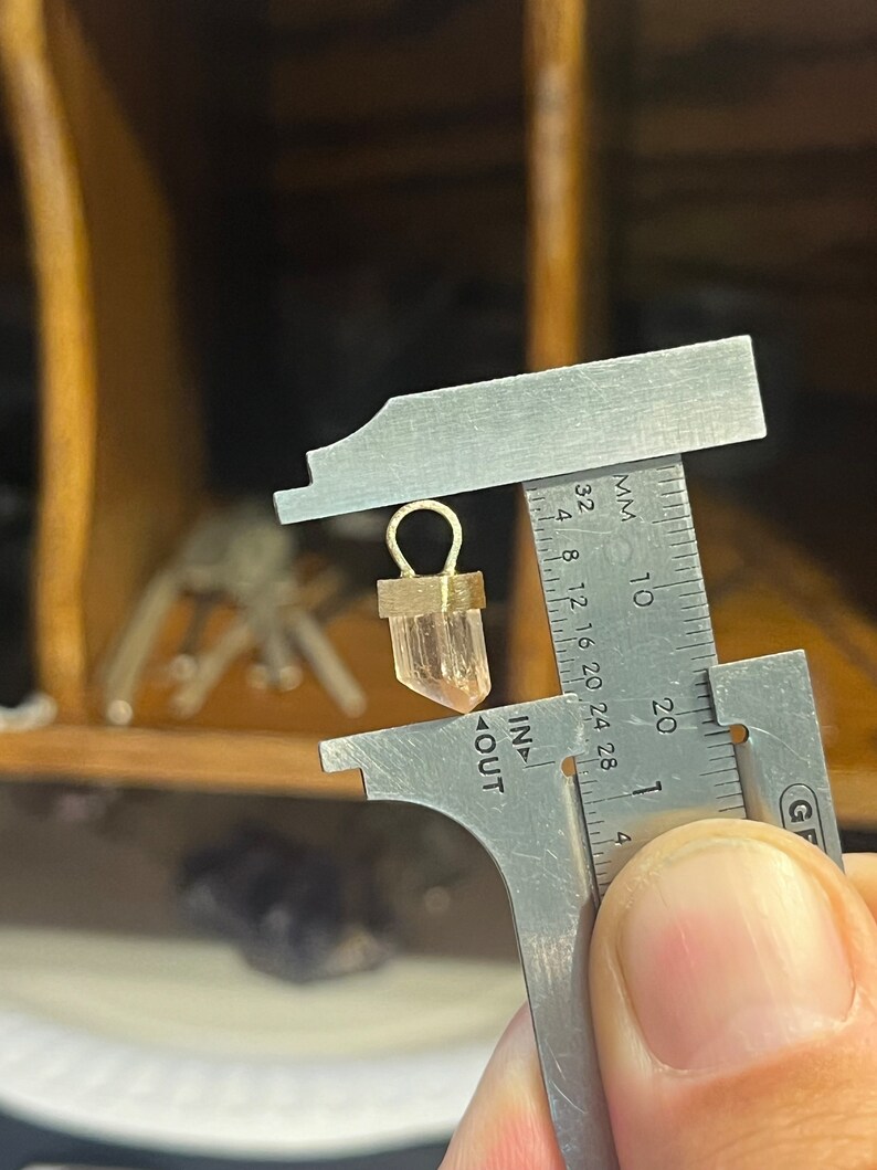 May include: A close-up of a clear quartz crystal pendant with a gold-toned setting. The pendant is being measured with a caliper, showing a measurement of approximately 1.5 centimeters.