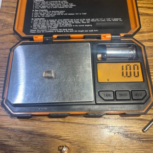 May include: A small, orange digital scale with a silver weighing platform. The scale is displaying 100 grams. The scale is sitting on a wooden surface. There is a small, gold-colored object on the surface next to the scale.