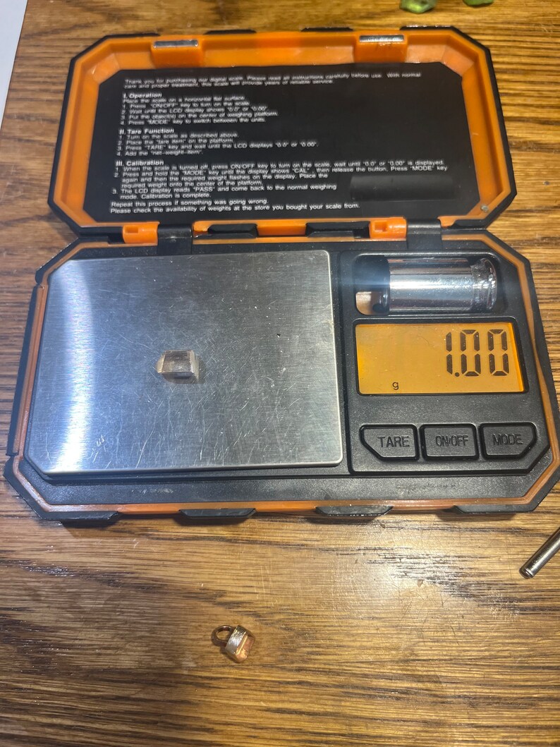 May include: A small, orange digital scale with a silver weighing platform. The scale is displaying 100 grams. The scale is sitting on a wooden surface. There is a small, gold-colored object on the surface next to the scale.